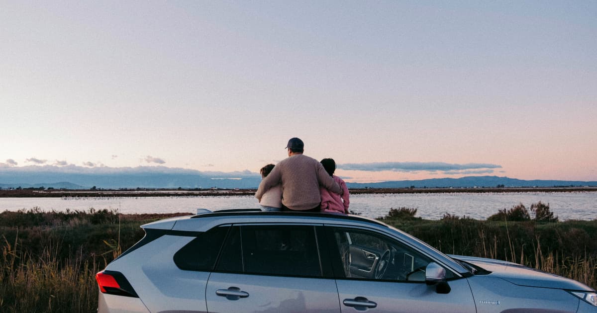 A family sitting on the roof of their compact SUV, watching the sunset over a lake